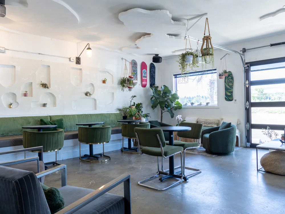 Tables and the long green banquette beside the roll-up garage door, open to the foothills, with hanging plants and skate decks above